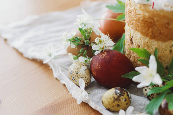 ハッピーイースター!スタイリッシュなイースターエッグ、春の花を咲かせ、素朴なテーブルの上に自家製イースターブレッド。テキスト用のスペース。現代の天然染色卵と伝統的なイースターフード - 写真・画像