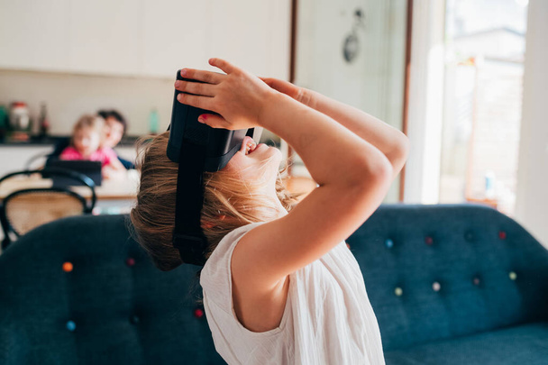 Enfant féminin à la maison en utilisant un casque de réalité virtuelle jouissant de jeux vidéo en réalité augmentée  - Photo, image
