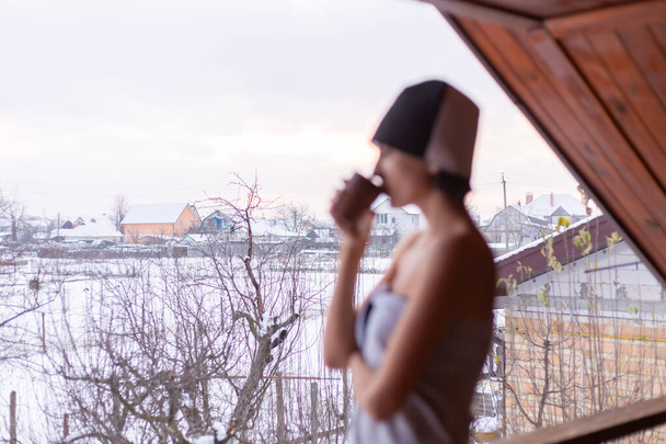 Junge Frau mit Mütze auf dem Kopf, in einem Handtuch, Tee in der Hand, steht im Winter auf dem Balkon - Foto, Bild