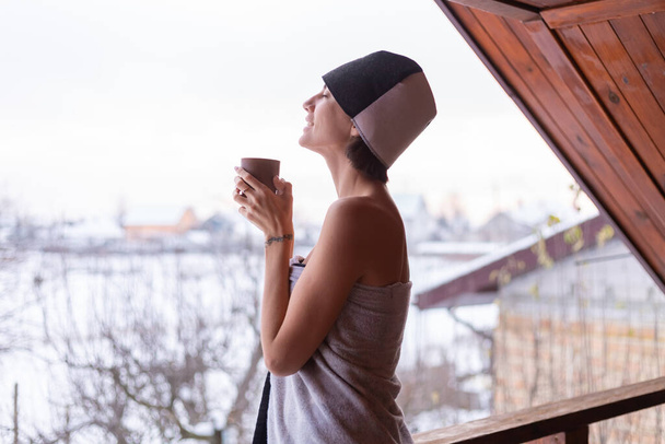 Junge Frau mit Mütze auf dem Kopf, in einem Handtuch, Tee in der Hand, steht im Winter auf dem Balkon - Foto, Bild