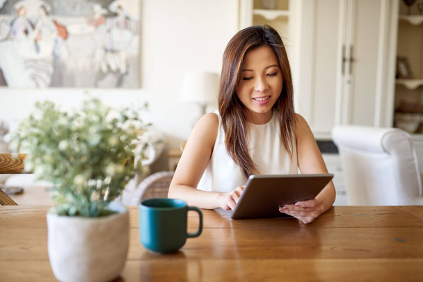 Sorridente giovane donna asiatica seduta da sola a un tavolo nel suo salotto a casa a lavorare online con un tablet digitale  - Foto, immagini