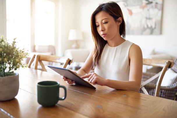 Jonge Aziatische vrouw zit aan een tafel in haar woonkamer thuis werken online met een digitale tablet  - Foto, afbeelding
