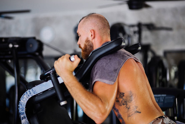 Fit Ευρωπαϊκό τατουάζ γενειοφόρος ισχυρός άνδρας των μυών κάνει καταλήψεις σε ένα εκπαιδευτικό μηχάνημα, αθλητικός εξοπλισμός φυσικής κατάστασης. - Φωτογραφία, εικόνα