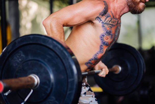 Un hombre fuerte tatuado con barba en el gimnasio. Muscular culturista chico haciendo ejercicios de barra - Foto, Imagen