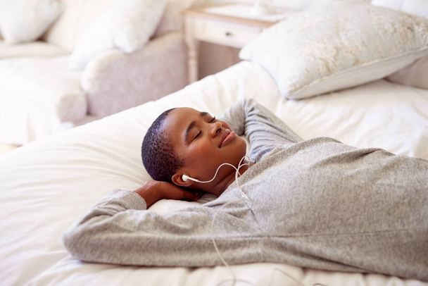 Улыбающаяся молодая афроамериканка расслабляется на кровати с руками за головой, слушая музыку в наушниках - Фото, изображение