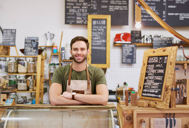 Portrait d'un beau jeune barista souriant et debout accueillant derrière le comptoir de commande d'un café branché  - Photo, image