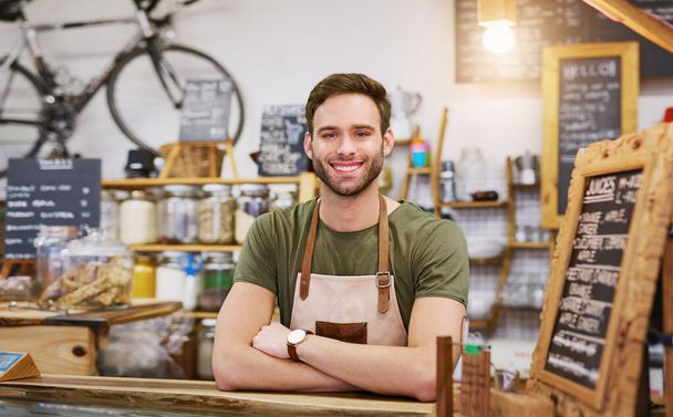 Portrét přátelského mladého baristy, který se usmívá, zatímco vřele stojí za přepážkou objednávek módní kavárny  - Fotografie, Obrázek