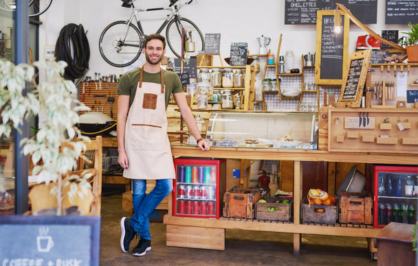 Retrato de um simpático jovem barista sorrindo enquanto está de pé acolhedor no balcão de encomendas de um café na moda  - Foto, Imagem