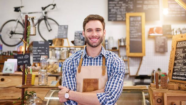 Portrait of a young barista smiling while standing with his arms crossed in a trendy cafe  - Photo, Image