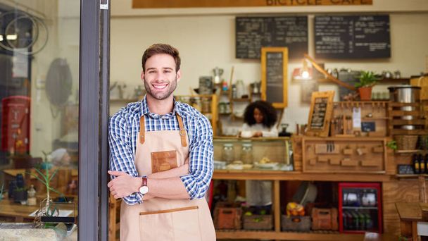 Portret młodego baristy uśmiechającego się i stojącego z skrzyżowanymi rękami, opierającego się na drzwiach modnej kawiarni  - Zdjęcie, obraz