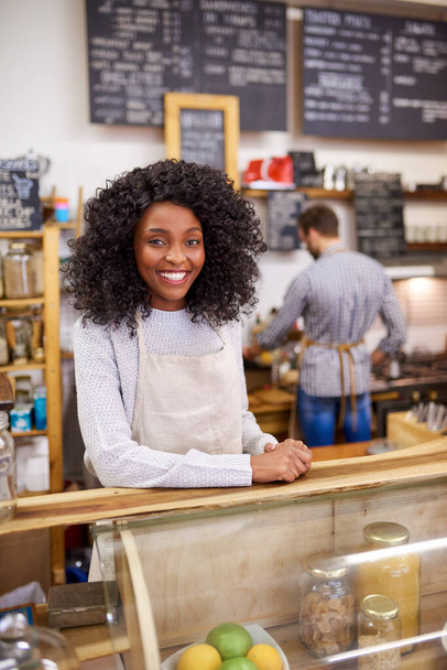 Egy mosolygó fiatal afro-amerikai barista portréja, aki egy trendi kávézó mögött áll egy munkatárssal a háttérben. - Fotó, kép