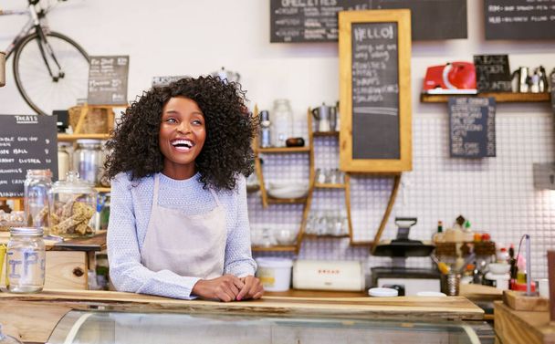 Φιλική νεαρή Αφρο-Αμερικανίδα μπαρίστα γελάει ενώ στέκεται πίσω από τον πάγκο ενός μοντέρνου καφέ - Φωτογραφία, εικόνα