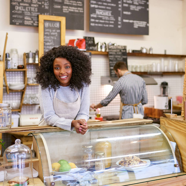 Retrato de un joven barista afroamericano sonriente parado detrás del mostrador de un café de moda con un colega trabajando detrás de ella - Foto, Imagen