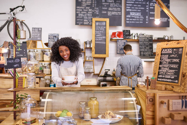 Egy mosolygó fiatal afro-amerikai barista portréja, aki felszolgál egy csésze friss cappuccinót, miközben egy trendi kávézó pultja mögött áll.  - Fotó, kép