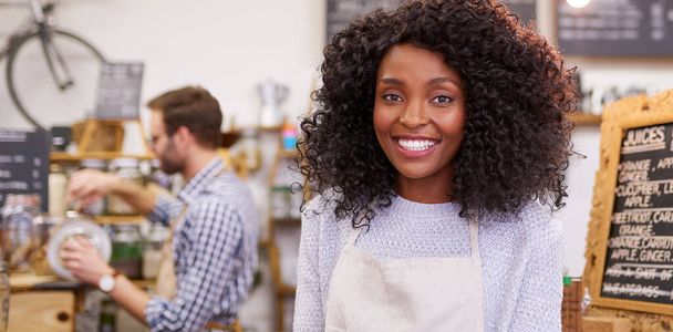 Portrait of a young African American barista smiling while standing in a trendy cafe with a coworker making coffee in the background  - Photo, Image