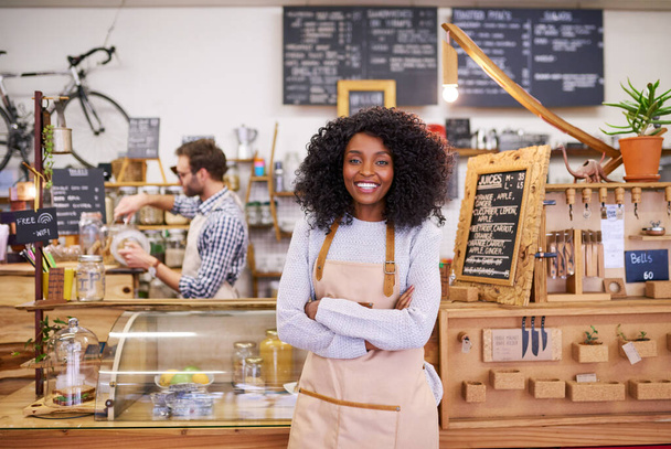 Portrait d'une jeune femme afro-américaine souriante debout les bras croisés dans un café branché avec un collègue en arrière-plan - Photo, image