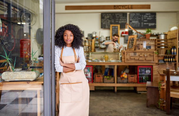 Retrato de una amigable joven barista afroamericana de pie con los brazos cruzados en la puerta de un café de moda  - Foto, Imagen
