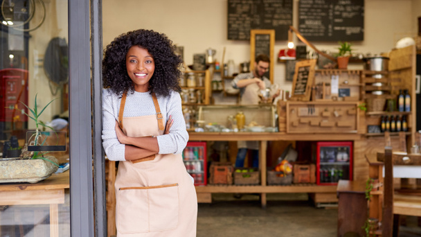 Portrait d'une jeune barista afro-américaine souriante penchée les bras croisés sur la porte d'un café branché  - Photo, image