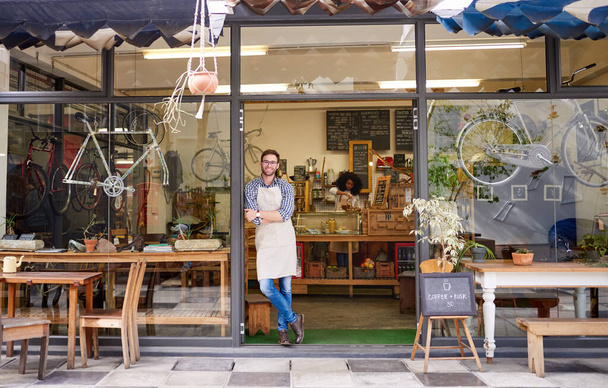 Portret van een knappe jonge barista glimlachend met zijn armen gekruist voor de deur van een trendy café  - Foto, afbeelding
