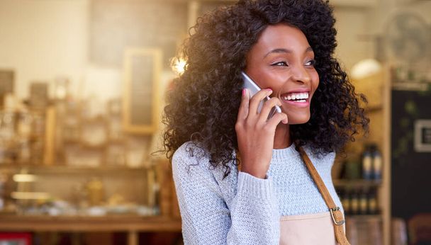 Sonriendo joven barista afroamericana hablando en su teléfono celular mientras está de pie en la entrada de un café de moda - Foto, imagen
