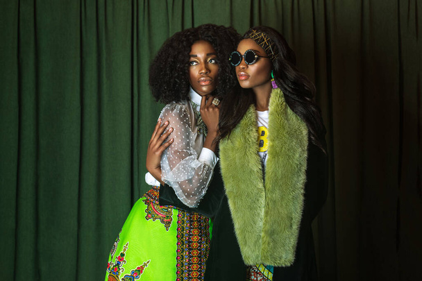 Retrato de 2 mujeres negras serenas con el pelo largo y rizado negro, hermoso maquillaje y labios suaves posando por sí mismas en un estudio con fondo de cortina verde usando ropa de diseñadores africanos. - Foto, imagen