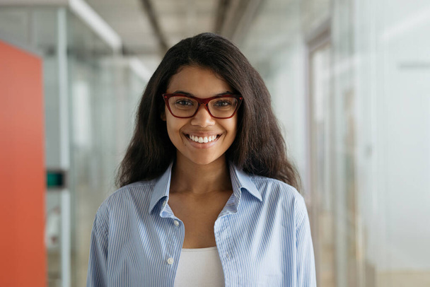 Mladá krásná Afroameričanka se stylovými brýlemi dívá do kamery a usmívá se. Portrét sebevědomého manažera stojící v moderní kanceláři   - Fotografie, Obrázek