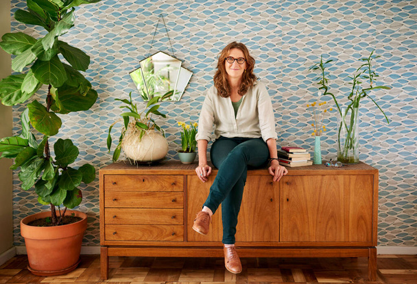 Портрет усміхненої зрілої жінки в окулярах, що сидить на шафі в її стильній квартирі
 - Фото, зображення