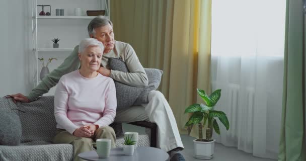 Pareja de ancianos caucásico anciano y mujer madura de pelo gris sentado abrazando abrazos en el sofá en el interior de la sala de estar moderna viendo la televisión hablando disfrutando pasar tiempo relajándose juntos en casa acogedora - Imágenes, Vídeo