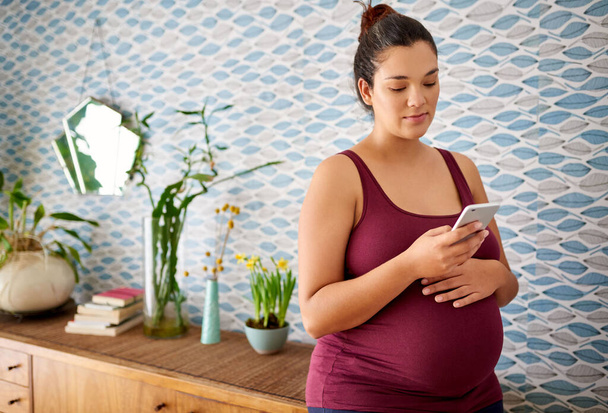 Mosolygó fiatal terhes nő áll a nappalijában otthon, és SMS-t olvas a mobilján. - Fotó, kép