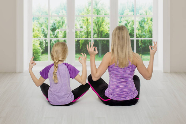  Aufnahme von Mutter und kleiner Tochter in ähnlichen Trainingsanzügen, die gemeinsam meditieren, während sie zu Hause auf dem Boden sitzen, Rückansicht - Foto, Bild