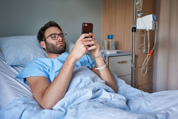 Młody człowiek leżący na szpitalnym łóżku i podłączony do kroplówki dożylnej za pomocą telefonu komórkowego       - Zdjęcie, obraz