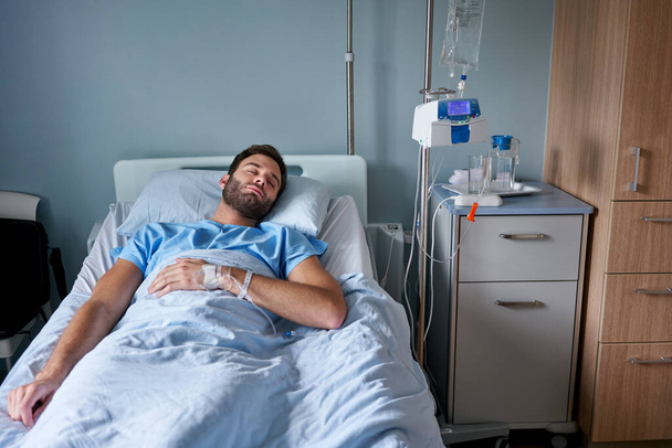 Молодой человек, прикрепленный к капельнице, крепко спит на кровати в больничной палате  - Фото, изображение