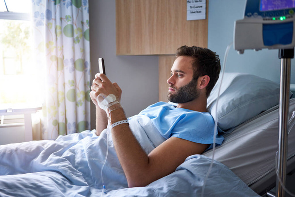 Genç bir adam, hastane odasındaki bir yatakta damardan damlayan bir damlaya bağlı olarak yatarken cep telefonu kullanıyor.                               - Fotoğraf, Görsel