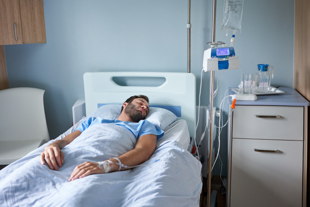 Jovem deitado dormindo em uma cama de hospital com um gotejamento intravenoso correndo em seu braço - Foto, Imagem