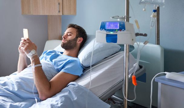 Młody mężczyzna leżący w szpitalnym łóżku, leczony kroplówką i korzystający z telefonu komórkowego. - Zdjęcie, obraz
