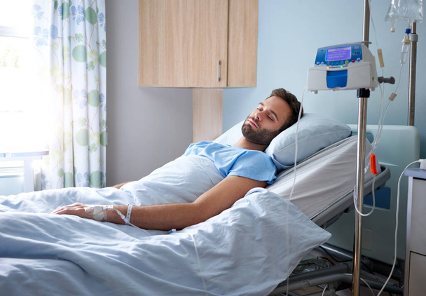 Хворий молодий чоловік, прикріплений до внутрішньовенної краплі, лежить на ліжку в лікарняній кімнаті
  - Фото, зображення