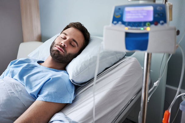 Больной молодой человек спит на кровати в больнице, получает внутривенное капельное лечение - Фото, изображение