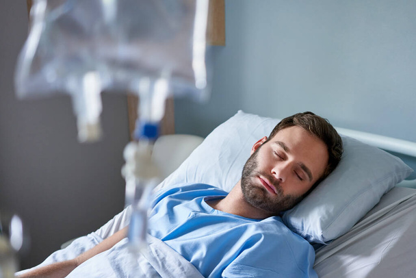 Больной молодой человек, спящий на кровати в больнице, получающий внутривенное капельное лечение - Фото, изображение