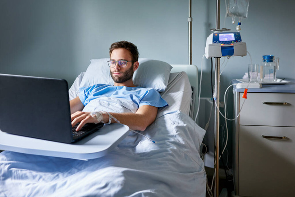 Młody mężczyzna przymocowany do kroplówki dożylnej, odpoczywający w szpitalu i pracujący na laptopie.   - Zdjęcie, obraz