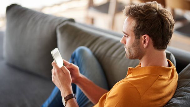 Giovane uomo sorridente che invia un messaggio sul suo cellulare mentre si rilassa da solo sul divano del suo soggiorno a casa - Foto, immagini