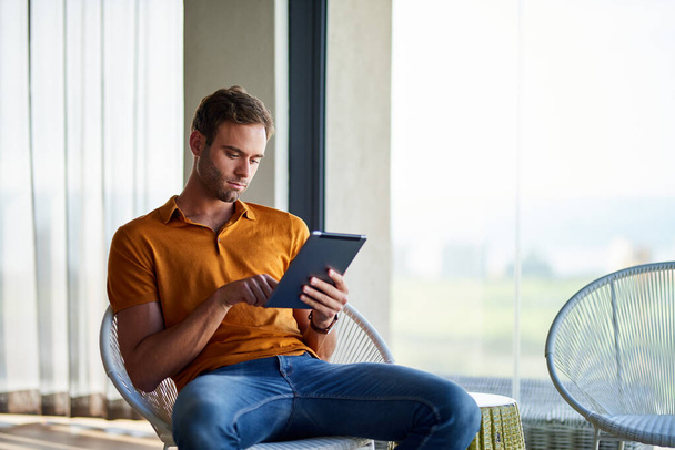 Giovane uomo che lavora online con un tablet digitale seduto da solo su una sedia nel suo appartamento moderno  - Foto, immagini