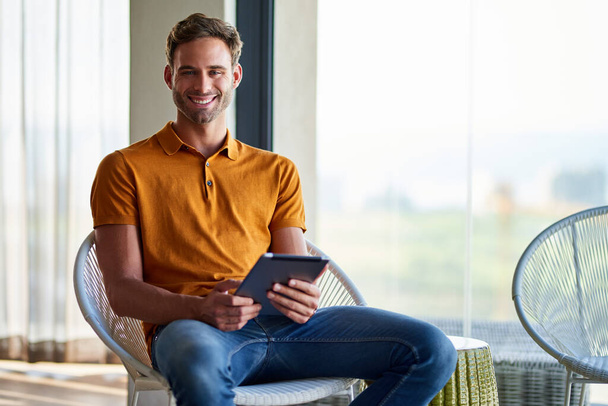 Πορτρέτο ενός χαμογελαστού νεαρού άνδρα που κάθεται μόνος του σε μια καρέκλα στο μοντέρνο διαμέρισμά του δουλεύοντας online με ένα ψηφιακό tablet - Φωτογραφία, εικόνα