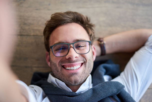 Υψηλής γωνίας πορτραίτο ενός χαμογελαστού νεαρού άνδρα που φορούσε γυαλιά και ένα πουλόβερ γύρω από τους ώμους του ξαπλωμένος στο πάτωμα βγάζοντας μια σέλφι - Φωτογραφία, εικόνα
