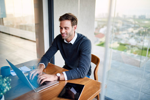 Glimlachende jonge zakenman zit aan tafel in zijn moderne appartement werken online met een laptop - Foto, afbeelding