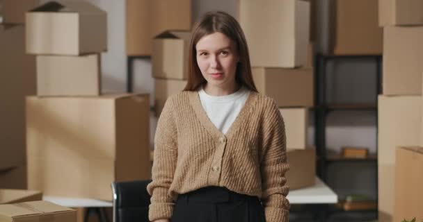 портрет молодой уверенной в себе предпринимательницы на складе с картонными коробками, смотрящей в камеру и складывающей руки - Кадры, видео