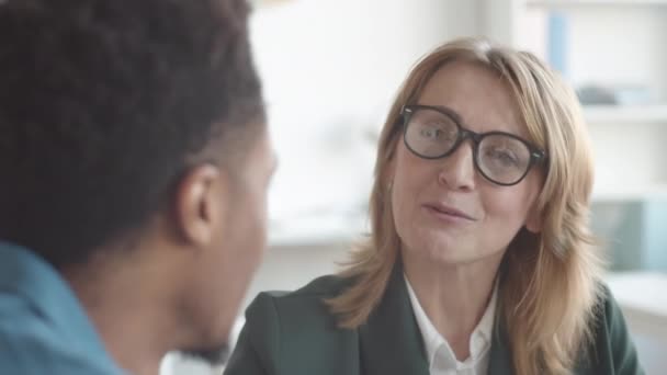 Zbliżenie zdjęcia uśmiechniętej białej kobiety w średnim wieku dyrektor HR w okularach i kurtce biznesowej rozmawiającej z młodszym współpracownikiem afro-amerykańskim podczas spotkania oceniającego w biurze - Materiał filmowy, wideo