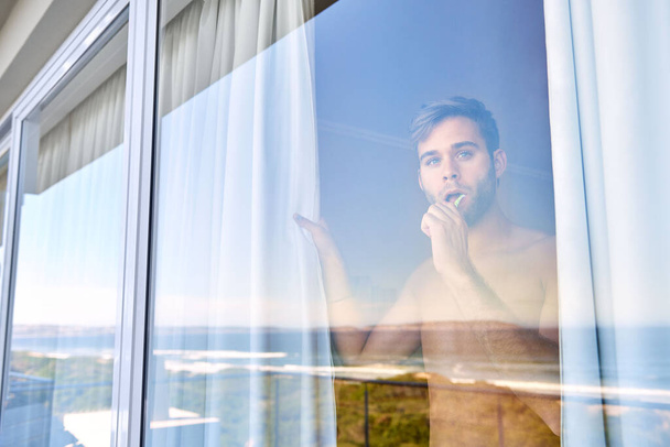 Blick durch das Glas eines gutaussehenden jungen Mannes, der sich beim Öffnen seines Vorhangs die Zähne putzt, um aus einem Fenster auf den Strand zu blicken - Foto, Bild