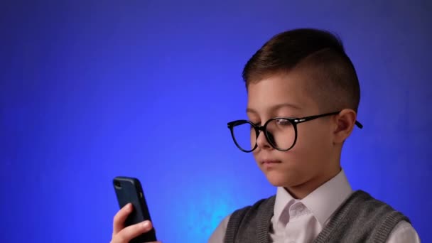 Menino bonito com óculos olha é surpreendido com telefone inteligente em fundo azul. Criança moderna é alegria olhando para gadget. - Filmagem, Vídeo