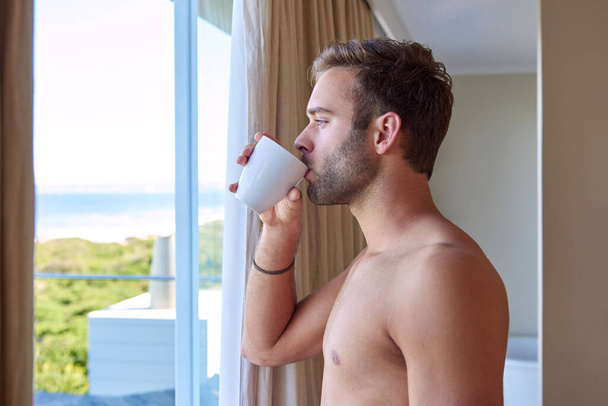 Komea nuori mies seisoo sisällä katsellen näkymää rannalle hotellin ikkunasta, siemaillen samalla ensimmäistä kupillista kahvia aamuksi. - Valokuva, kuva