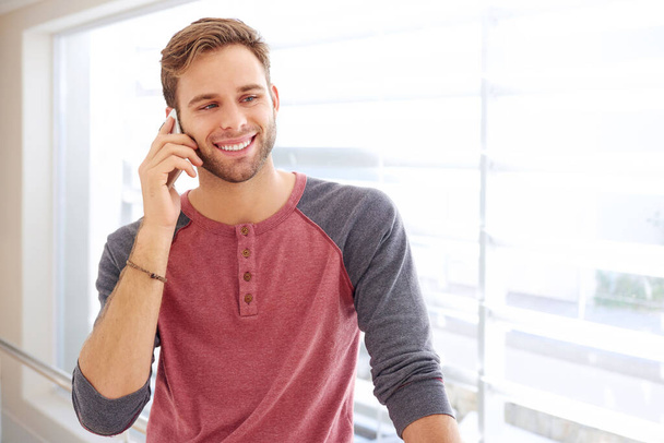 Όμορφος νεαρός άνδρας με μοντέρνα casual ρούχα χαμογελά και κοιτάζει αλλού ενώ έχει μια συνομιλία στο κινητό του τηλέφωνο σε εσωτερικούς χώρους - Φωτογραφία, εικόνα